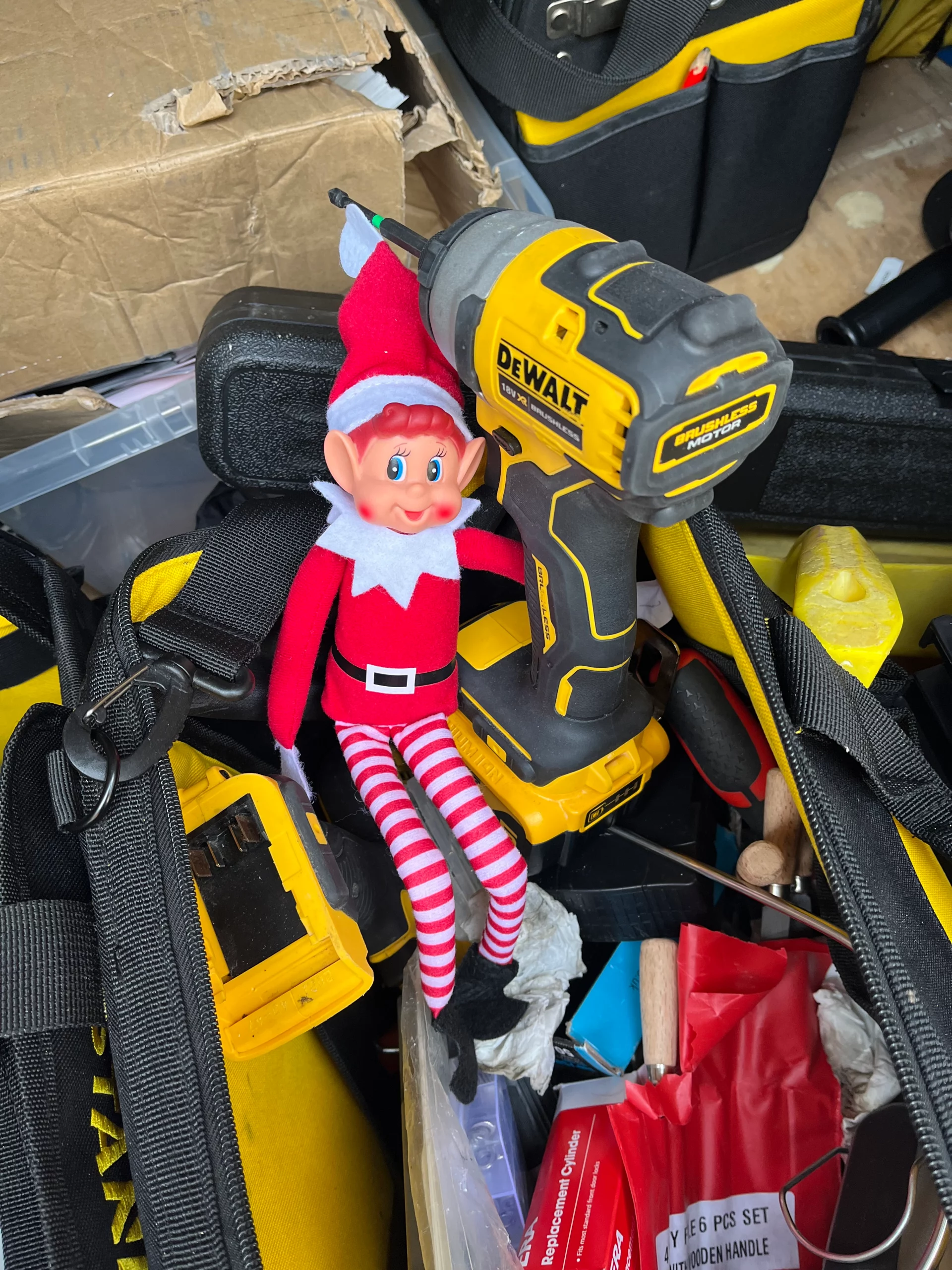 Smithy the Elf sitting on a yellow DeWalt drill among locksmith tools and equipment, spreading festive cheer with his signature striped outfit and cheeky grin.