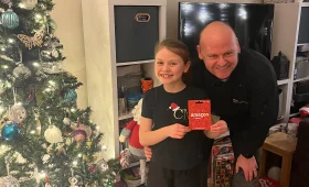 James from Key Objective Locksmiths presenting a £75 Amazon voucher to the McKeown family in Chichester. The festive setting includes a decorated Christmas tree and wrapped gifts.