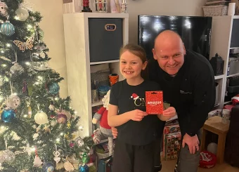 James from Key Objective Locksmiths presenting a £75 Amazon voucher to the McKeown family in Chichester. The festive setting includes a decorated Christmas tree and wrapped gifts.