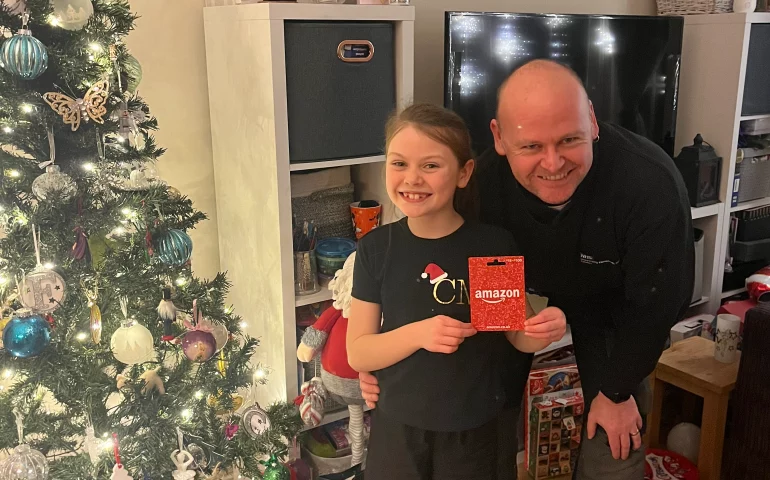 James from Key Objective Locksmiths presenting a £75 Amazon voucher to the McKeown family in Chichester. The festive setting includes a decorated Christmas tree and wrapped gifts.
