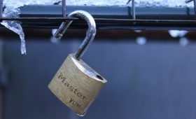 A brass Master padlock with its shackle unlocked, hanging on a metal wire fence covered in frost and icicles.