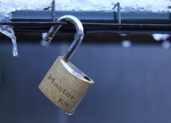 A brass Master padlock with its shackle unlocked, hanging on a metal wire fence covered in frost and icicles.