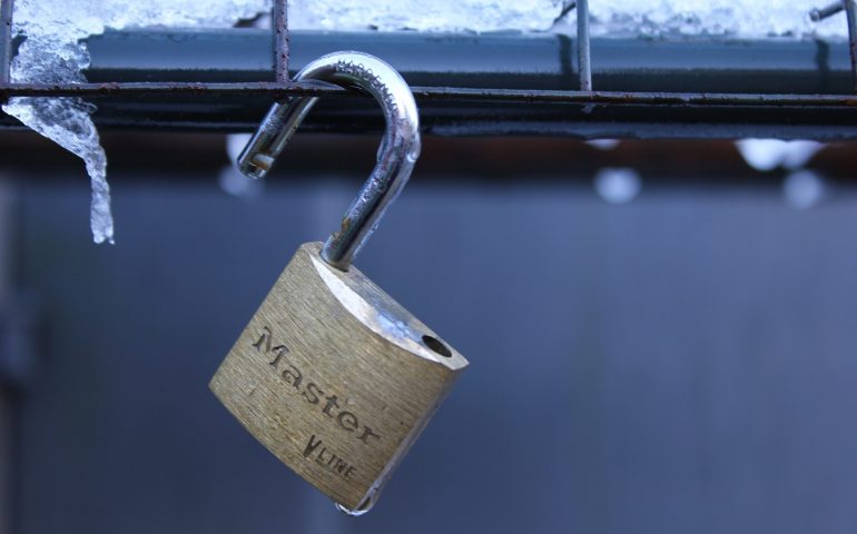 A brass Master padlock with its shackle unlocked, hanging on a metal wire fence covered in frost and icicles.